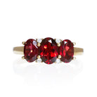 A product photo of a Garnet Trio & Diamond Ring in Yellow Gold sitting on a plain white background. The largest garnet centrestone measures 7x5mm, with 4 tiny diamonds nestled against it, two on each side. The garnet stones are deep red, reflecting sanguine hues across their multi-faceted edges.