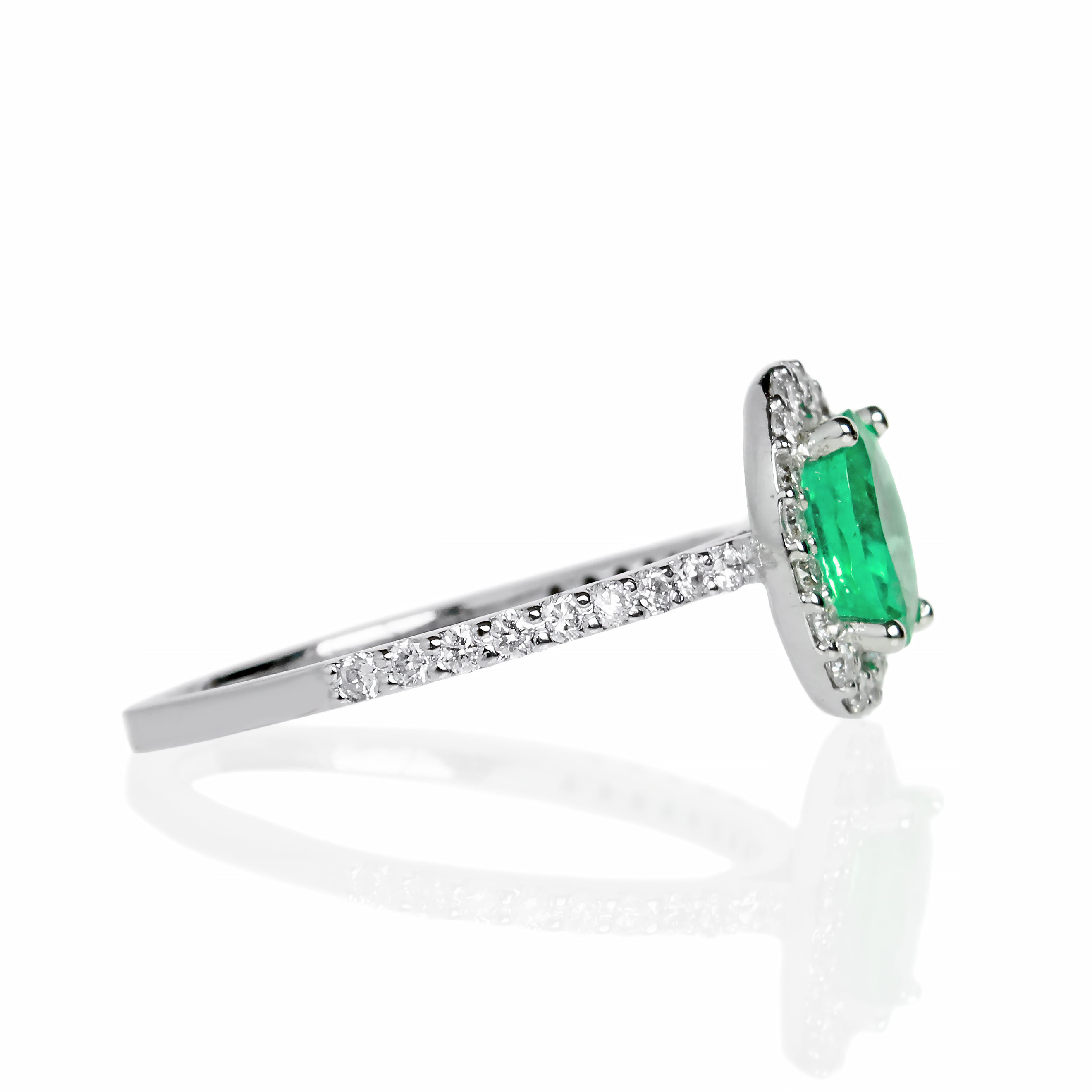 A product photo of a fine emerald ring in 14k white gold on a white background. A large, brilliant oval jewel in the centre reflects mint-coloured light, and is framed by a ring of white diamonds. A simple round band continues the diamond detailing for half of its length, smoothing out into a simple white gold back.