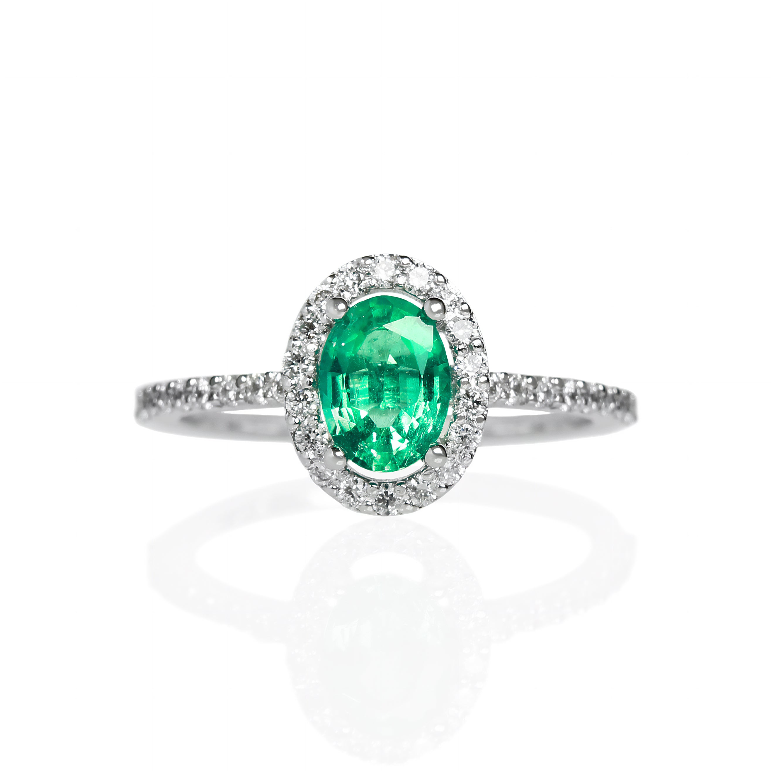 A product photo of a fine emerald ring in 14k white gold on a white background. A large, brilliant oval jewel in the centre reflects mint-coloured light, and is framed by a ring of white diamonds. A simple round band continues the diamond detailing for half of its length, smoothing out into a simple white gold back.