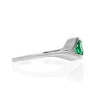 A product photo of a white gold emerald ring sitting on a white background. The band splits and curves around the single, round emerald centre stone, holding it in place like an eye. The emerald stone reflects minty shades of light from its many edges.