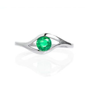A product photo of a white gold emerald ring sitting on a white background. The band splits and curves around the single, round emerald centre stone, holding it in place like an eye. The emerald stone reflects minty shades of light from its many edges.