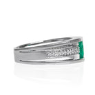 A product photo of a white gold trio-banded ring with diamond detailing and a natural emerald centre stone sitting on a white background. The top and bottom bands are smooth and minimalistic, hugging the top and bottom of the oval emerald centre stone, whilst the middle band (meeting on either side of the stone) is bejewelled with many tiny diamonds.