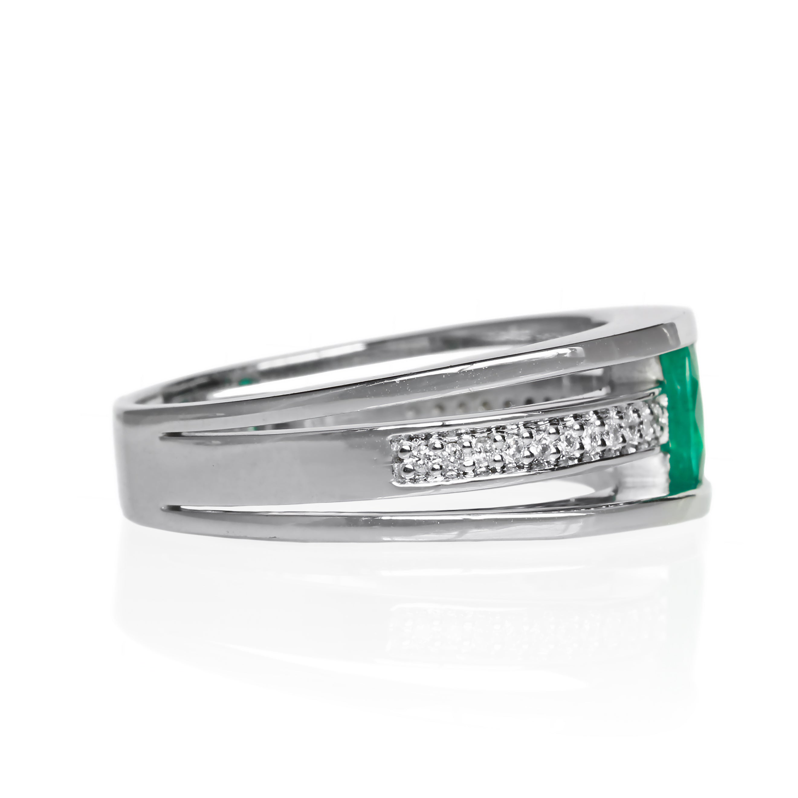 A product photo of a white gold trio-banded ring with diamond detailing and a natural emerald centre stone sitting on a white background. The top and bottom bands are smooth and minimalistic, hugging the top and bottom of the oval emerald centre stone, whilst the middle band (meeting on either side of the stone) is bejewelled with many tiny diamonds.