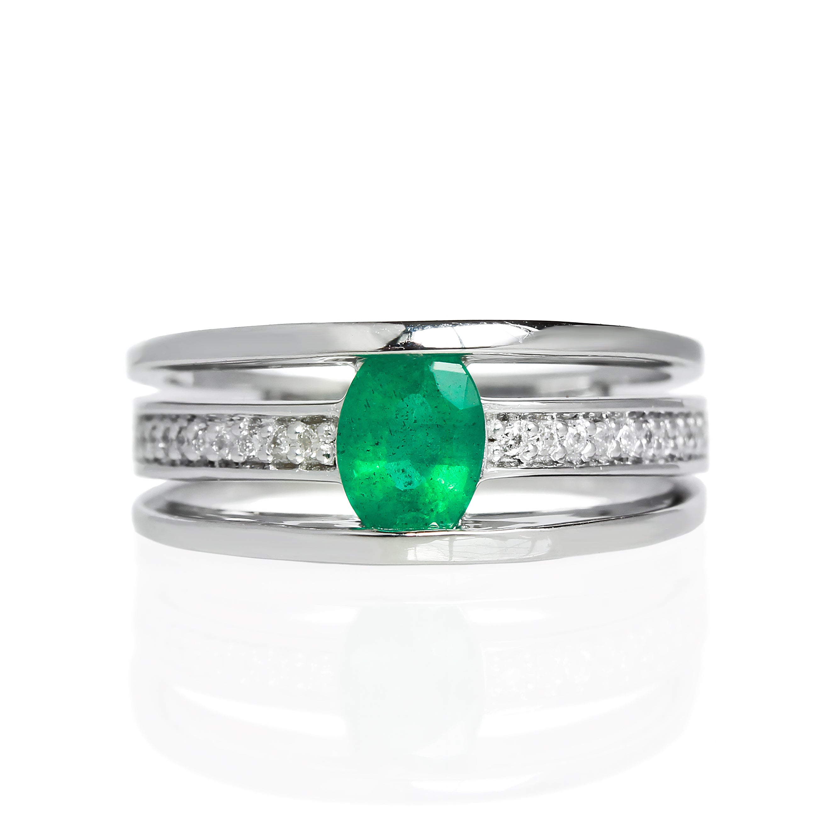 A product photo of a white gold trio-banded ring with diamond detailing and a natural emerald centre stone sitting on a white background. The top and bottom bands are smooth and minimalistic, hugging the top and bottom of the oval emerald centre stone, whilst the middle band (meeting on either side of the stone) is bejewelled with many tiny diamonds.