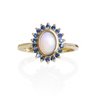 A product photo of a vintage styled white opal ring in 9ct yellow gold on a white background. A large, fiery oval stone in the centre reflects multi-coloured light, and is framed by a ring of blue sapphires. The band is simple and rounded.