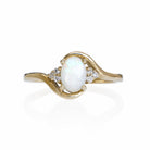 A product photo of a mystical white opal and diamond fantasy ring in solid 9k yellow gold on a white background. The fiery white opal is held in place by 4 claws and reflects light off of its many facets, while 3 diamonds sit on either side.. The smooth yellow gold band curves elegantly, meeting at the top and bottom of the opal and diamond arrangement.