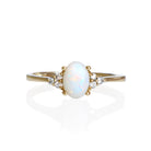 A product photo of a yellow gold white opal and diamond trio ring sitting on a white background. The oval opal gemstone stands in stark contrast to the little clusters of three white diamond stones on either side.