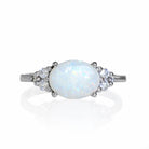 A product photo of a white gold white opal and diamond trio ring sitting on a white background. The large oval opal gemstone stands in stark contrast to the little clusters of three white diamond stones on either side.