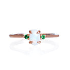 A product photo of a delicate 9ct rose gold 6x4mm white opal and tsavorite trio ring sitting on a white background. The fiery opal centre stone is hugged on either side by a bright green tsavorite jewel.