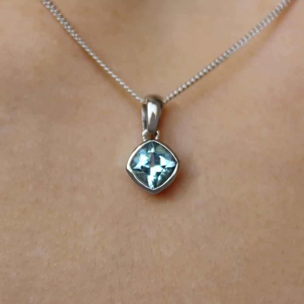 A product photo of a white gold aquamarine pendant suspended by a chain against a white background. The squared cushion-cut stone is oriented diagonally and is encased in a thick layer of white gold bezel setting. It reflects pale baby blue colours from its many edges.