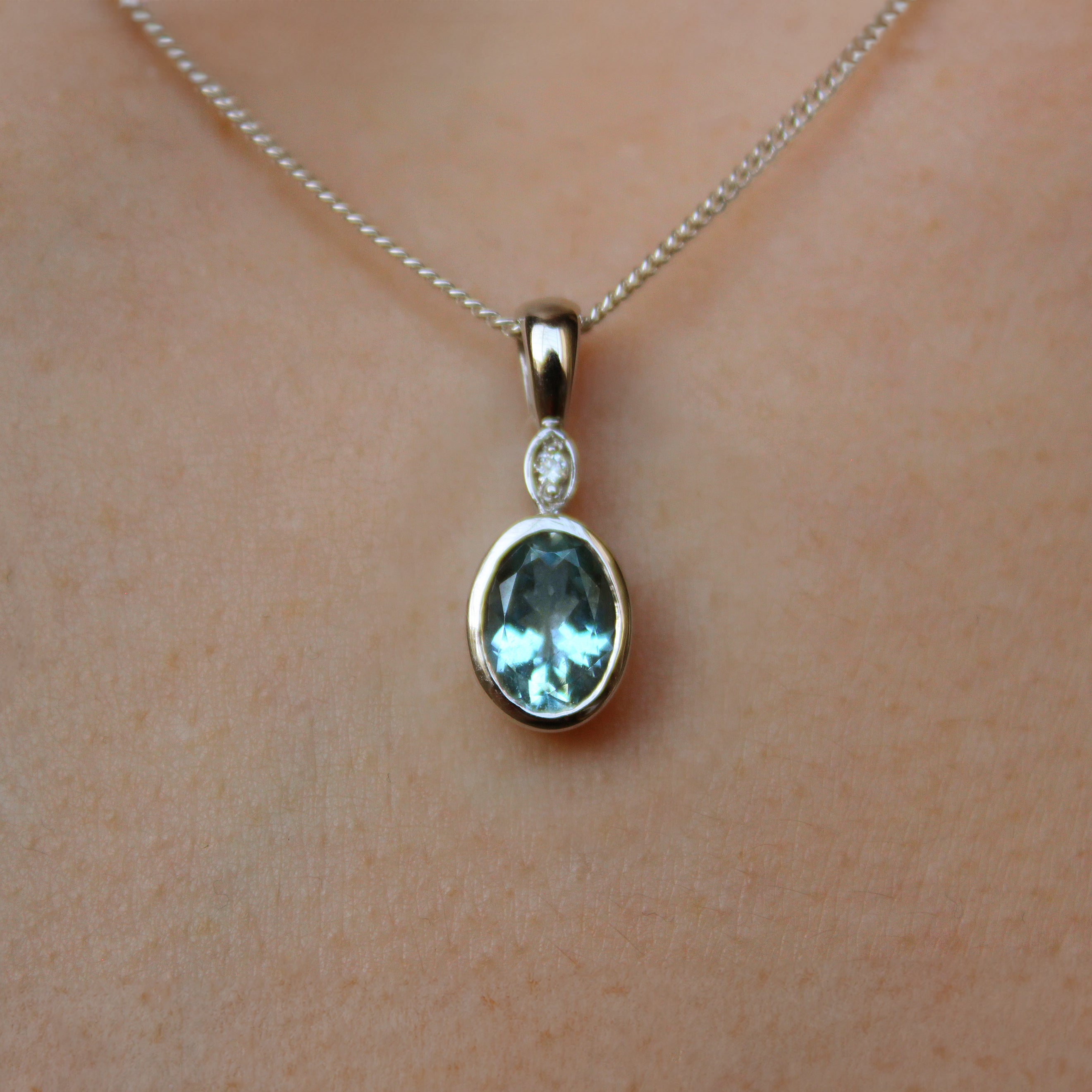 A product photo of a Bezel-set Aquamarine Pendant and diamond in 9ct White Gold suspended by a gold chain on a plain white background. Above the bezel-set aquamarine stone sits a single white diamond, also encased in white gold.