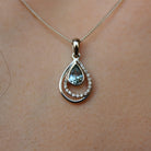 A product photo of a modern and elegant aquamarine and diamond pendant in 9k white gold. The pear-shaped bezel--set aquamarine jewel sits at the base of a teardrop-shaped frame of solid white gold, adorned with diamond details, which is echoed by a another teardrop-shaped smooth golden frame slightly offset from the centre.