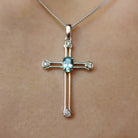 A product photo of a white gold aquamarine and diamond pendant in the shape of a Christian cross. At the ends of each limb, a tiny white diamond marks the tip, to a total of 4 diamonds. At the heart of the cross is a dainty baby blue aquamarine centre stone.