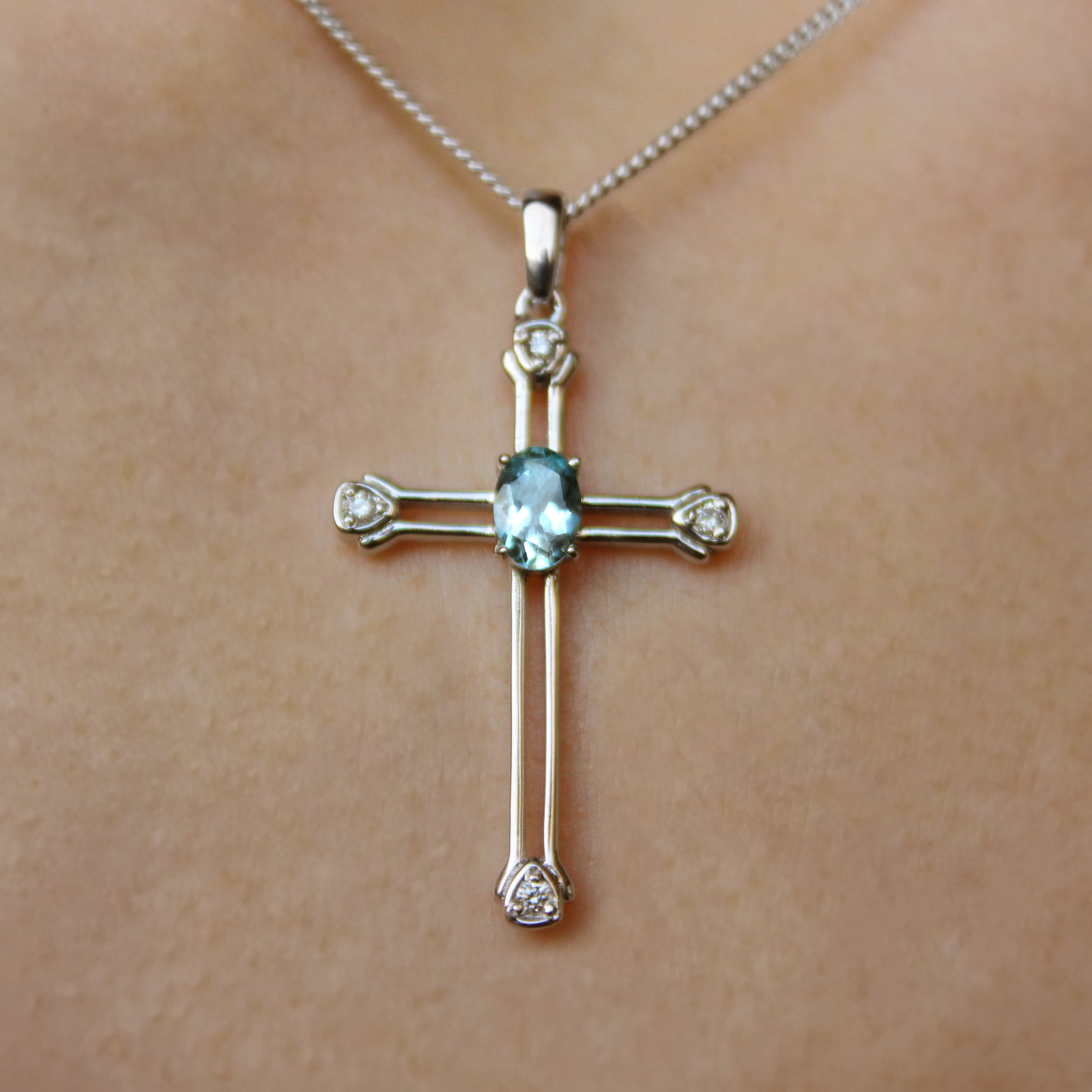 A product photo of a white gold aquamarine and diamond pendant in the shape of a Christian cross. At the ends of each limb, a tiny white diamond marks the tip, to a total of 4 diamonds. At the heart of the cross is a dainty baby blue aquamarine centre stone.