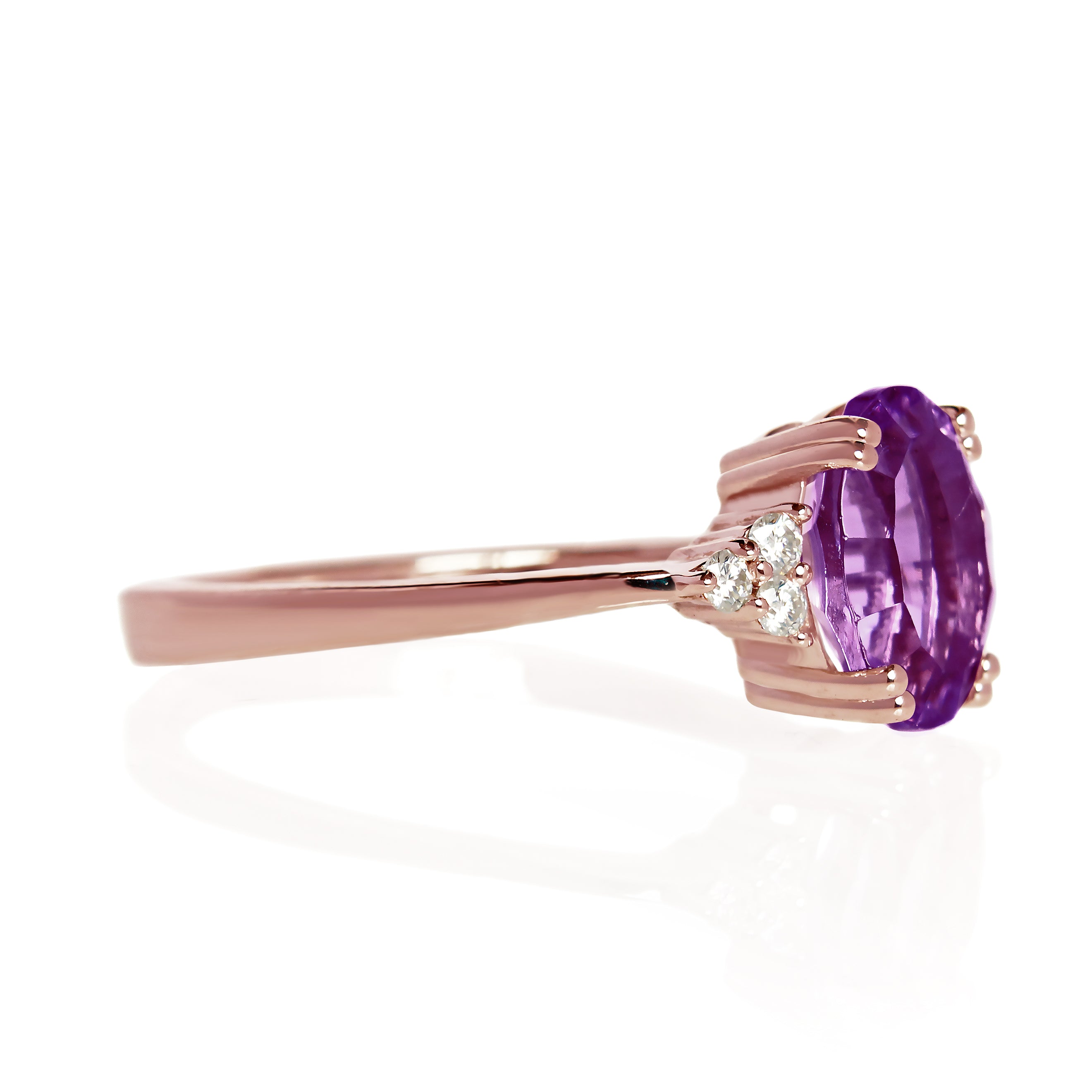 A product photo of a rose gold amethyst and diamond trio ring sitting on a white background. The oval purple amethyst gemstone stands in stark contrast to the little clusters of three white diamond stones on either side.