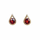 A product photo of 9ct yellow gold garnet stud earrings sitting on a white background. The round stones are encased by thick, yellow gold frames in the shapes of teardrops. The deep red garnet stones reflect sanguine light from their many edges.