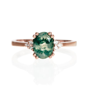 A product photo of a rose gold moss agate and diamond trio ring sitting on a white background. The oval, naturally-included moss agate gemstone stands in stark contrast to the little clusters of three classic white diamond stones on either side.