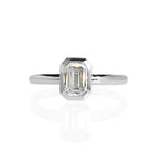 A product photo of a 14 karat white gold lab diamond bezel solitaire engagement ring sitting in the sun on a white textured background. The brilliant, colourless 0.72ct emerald-cut diamond measures about 6.3mm lengthwise, and is held in place by a delicate golden-framed bezel setting.