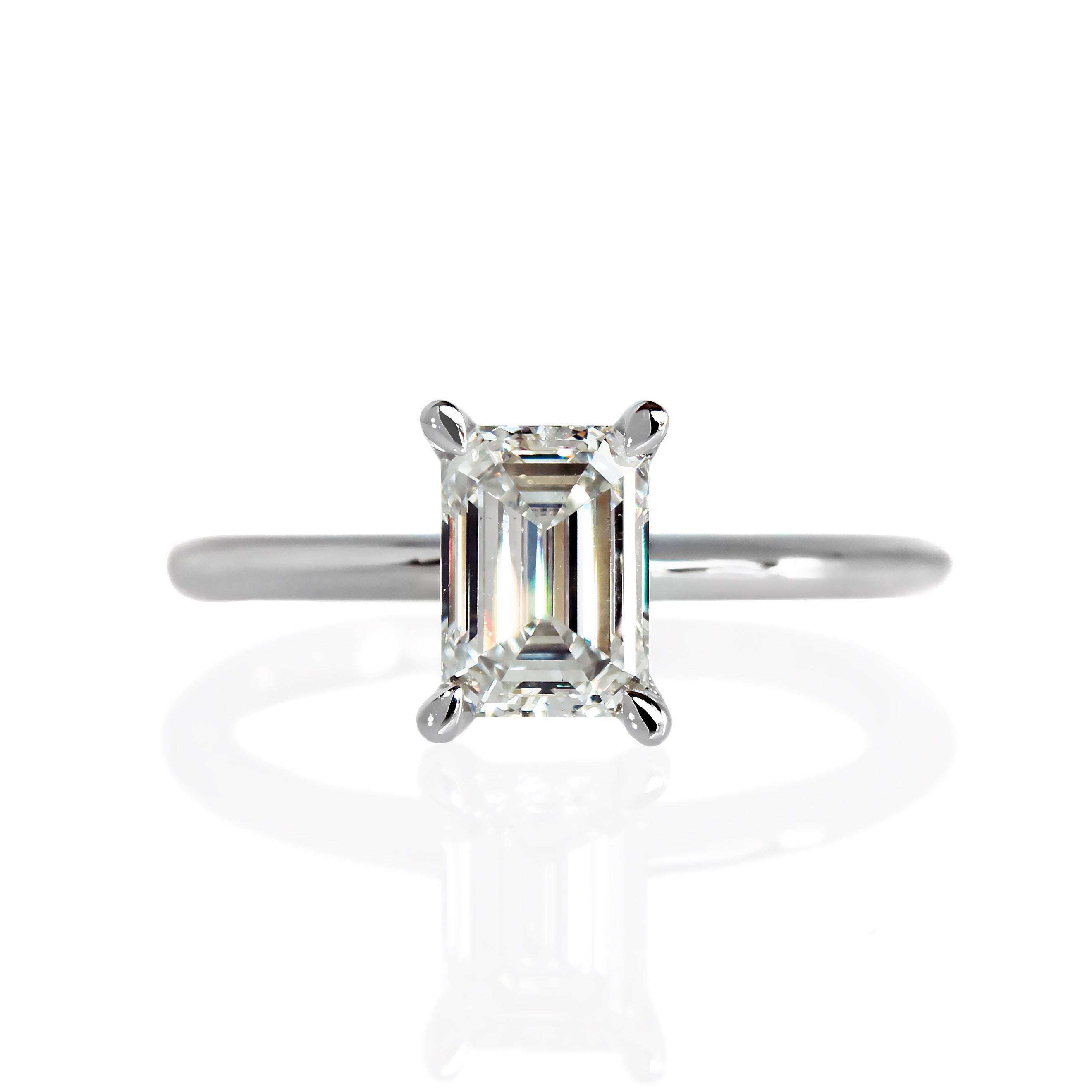 A product photo of a 14 karat white gold lab diamond engagement ring sitting in the sun on a white textured background. The brilliant, colourless 1ct emerald-cut diamond measures about 6.7mm lengthwise, and is held in place by 4 delicate golden claws in a prong setting.