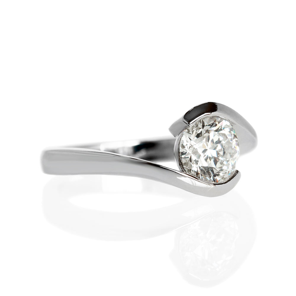 A product photo of a 14 karat white gold lab diamond solitaire engagement ring sitting in the sun on a white textured background. The brilliant, colourless 1 carat round-cut diamond measures 6.3mm lengthwise, and is held in place by a thick white gold band that curves around the top and bottom of the stone in a tension setting.
