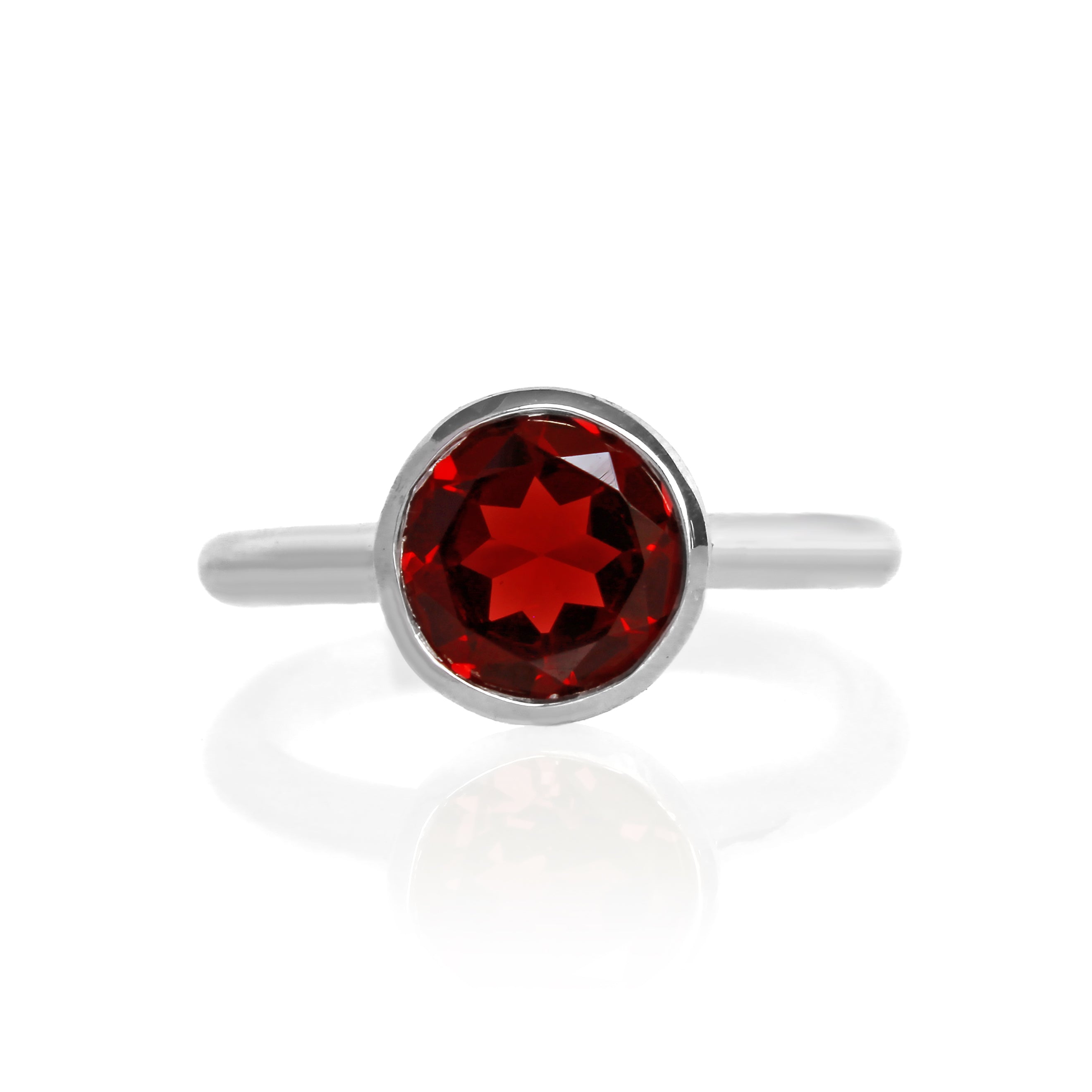 A product photo of a silver ring with a bezel-set garnet centre stone sitting on a white background. The silver band is simple and smooth, connecting on either side of a large, statement-sized garnet stone surrounded by a solid frame of silver.