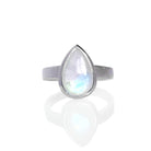 A product photo of a silver moonstone ring on a white background. The band is tall and thick. The 12x8mm pear cabochon moonstone has a blue/purple sheen, with unique milky natural inclusions. The stone is set in a thick silver bezel setting. 