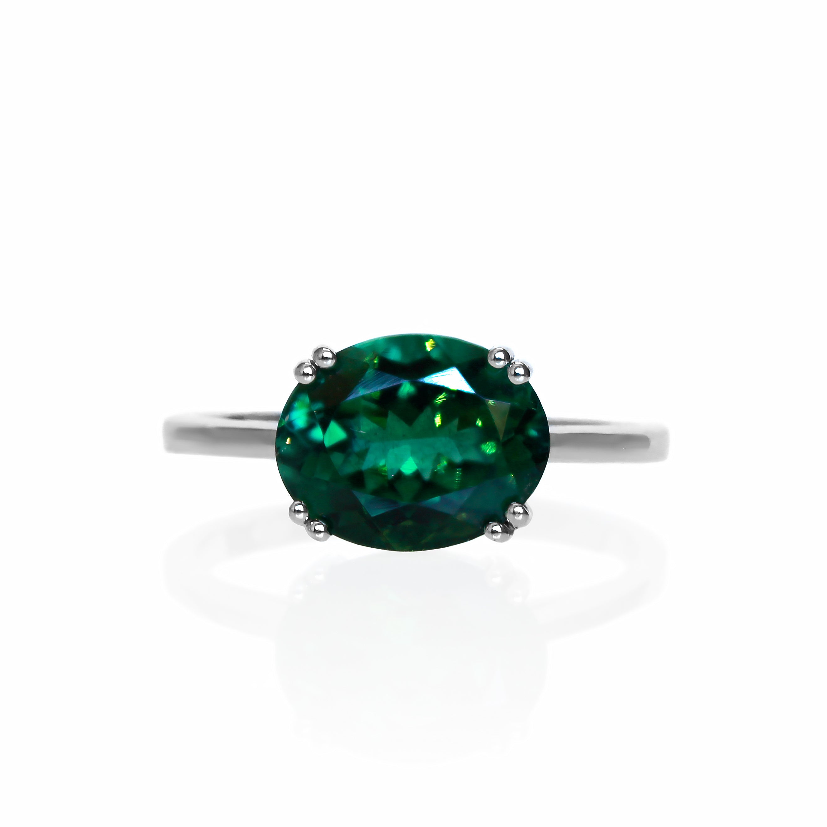 A product photo of a white gold ring with an impressive oval dark green tourmaline centre stone sitting on a white background. The band is simple and rounded, with a single 10x8mm oval, faceted deep green tourmaline centre stone, held in place with 8 delicate golden claws, each split into pairs of 2.