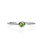 A product photo of a delicate white gold stacking ring with a tiny, bezel-set peridot in the centre sitting on a white background. The band is slim and thread-like, with the focus drawn to the petite 3mm glinting green centre stone.