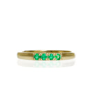 A product photo of a boldly minimalist wedding band made of solid 9k yellow gold sitting on a white background. The front is embedded with 4 tiny gemstones, all a delicate emerald green.