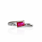 A product photo of an asymmetric ruby ring in 9 karat white gold on a white background. The smooth golden band is split, one side curving up and the other down to meet at the top and bottom of the horizontally-oriented pink ruby stone in an asymmetrical fashion.