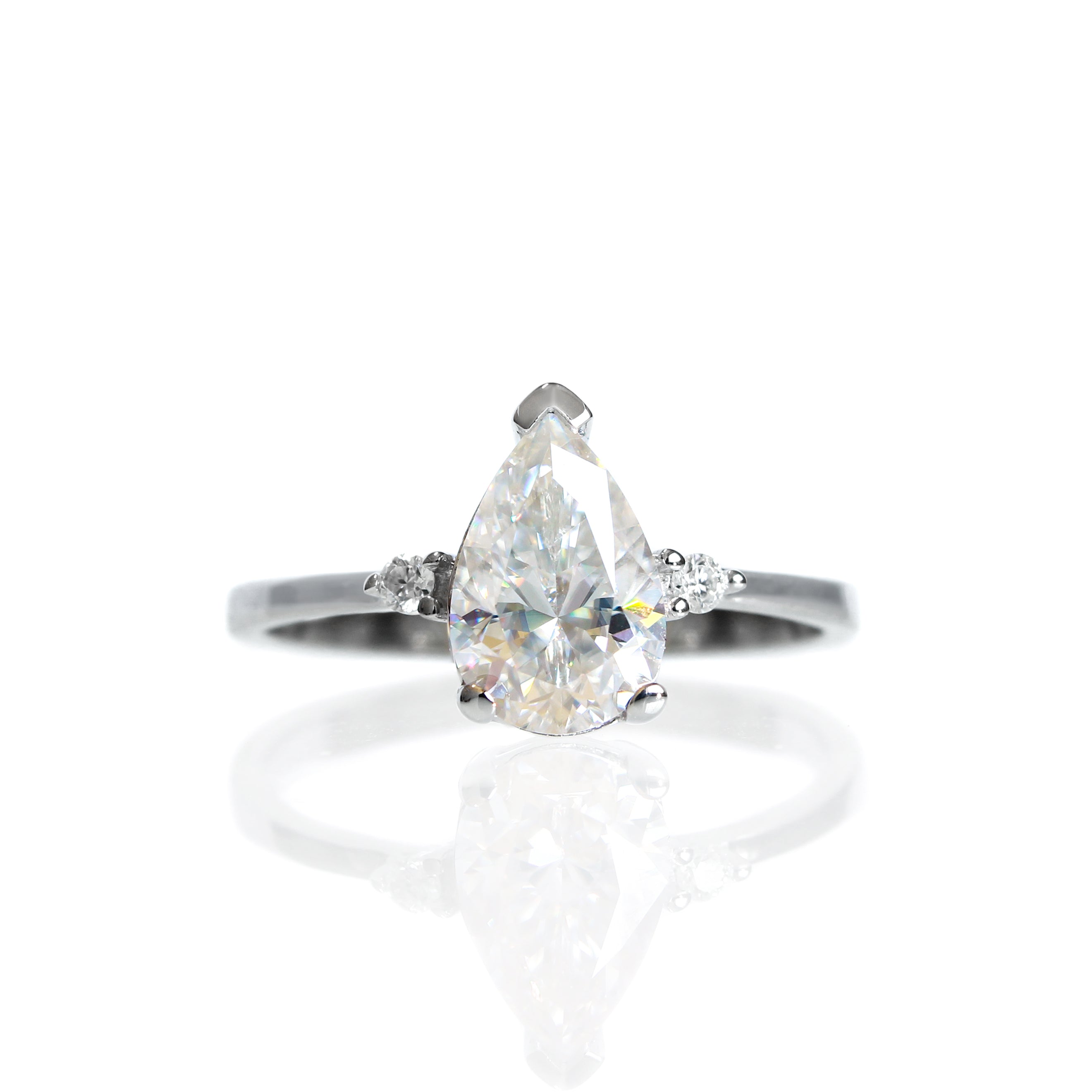 A product photo of a 9 karat white gold ring with a pear-cut clear moissanite centre stone sitting on a white background. The golden band is simple and smooth, meeting elegantly on either side of the moissanite centre stone, hugged by a single white moissanite on either side.