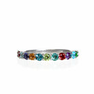 A silver multi-gemstone eternity ring sitting on a white reflective background. The ring is made up of 13 2.5mm round-cut semi-precious gemstones in a rainbow of colours.