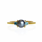 A product photo of a solid 9 karat yellow gold pearl engagement ring sitting on a white background. The pearl is a 5mm perfectly spherical steel grey pearl, hugged on either side by a delicate aquamarine jewel.