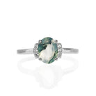 A product photo of a silver moss agate and moissanite trio ring sitting on a white background. The oval, naturally-included moss agate gemstone stands in stark contrast to the little clusters of three classic white moissanite stones on either side.