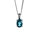 A product photo of a silver London Blue topaz pendant suspended by a silver chain against a white background. The oval gemstone is held in place by 4 small claws, and reflects deep blue hues from its many edges