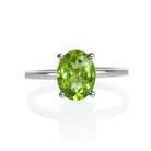 A product photo of an oval silver solitaire ring on a white background. The band is rounded and smooth, and the 9x7mm faceted peridot gemstone, glinting chartreuse in the light, is held in place by 4 delicate silver claws.