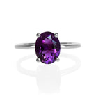 A product photo of an oval silver solitaire ring on a white background. The band is rounded and smooth, and the 9x7mm faceted amethyst gemstone, glinting deep purple in the light, is held in place by 4 delicate silver claws.