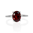 A product photo of an oval silver solitaire ring on a white background. The band is rounded and smooth, and the 9x7mm faceted garnet, glinting crimson in the light, is held in place by 4 delicate silver claws.