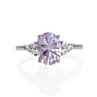 A product photo of a silver pink amethyst and moissanite trio engagement ring sitting on a white background. The unusual cut and colouring of the oval, laser-cut pink amethyst gemstone stands in stark contrast to the little clusters of three classic white moissanite stones on either side.