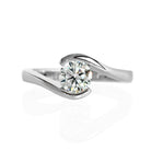 A product photo of a 14 karat white gold lab diamond solitaire engagement ring sitting in the sun on a white textured background. The brilliant, colourless 0.70 carat round-cut diamond measures 5mm lengthwise, and is held in place by a thick white gold band that curves around the top and bottom of the stone in a tension setting.