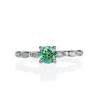 A product photo of a mint green colour moissanite engagement ring on a white background. The ring consists of a single 5mm round mint green moissanite centre stone, reflecting rainbow colours in the light, and 4 0.01ct white moissanite details studded along its filigree-detailed silver band.