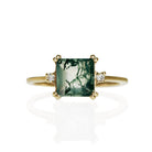 A product photo of a 7x7mm square cut moss agate and diamond ring on a white background. The ring is made of solid 9 karat yellow gold, with a smooth band and double-clawed settings on each corner of the moss agate centre stone - holding it in place. On either side of the stone is a 0.02ct natural diamond, held in place by 4 delicate golden prongs.