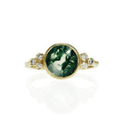 A product photo of a 8mm round cut moss agate and diamond ring on a white background. The ring is made of solid 9 karat yellow gold, with a smooth band. A thick frame of gold holds the moss agate centre stone and diamond details in place.