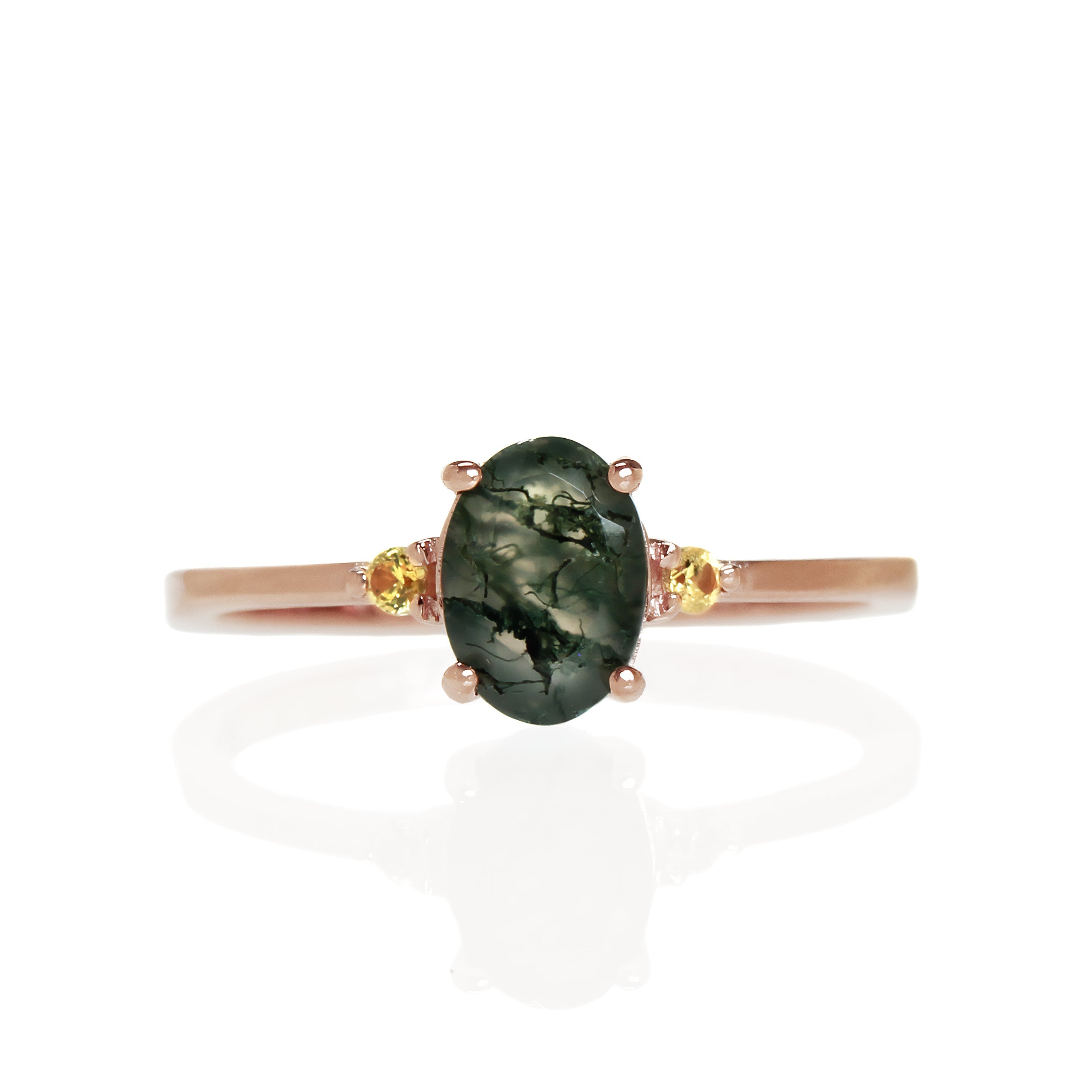 A product photo of a rose gold moss agate ring sitting against a white background. The rose gold band is plain and smooth, and the centre oval-cut dark green moss agate quartz stone is framed by a single yellow sapphire jewel on either side.