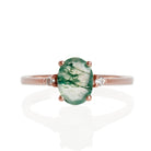 A product photo of a rose gold moss agate and diamond ring sitting against a white background. The rose gold band is plain and smooth, and the centre oval-cut light moss agate stone is framed by a single white diamond on either side.