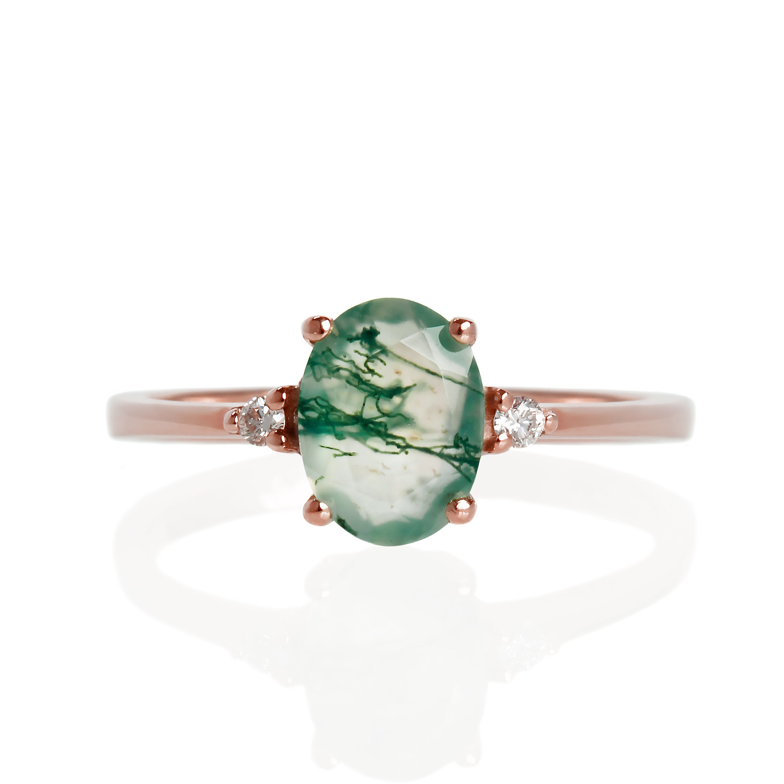 A product photo of a rose gold moss agate and diamond ring sitting against a white background. The rose gold band is plain and smooth, and the centre oval-cut light moss agate stone is framed by a single white diamond on either side.