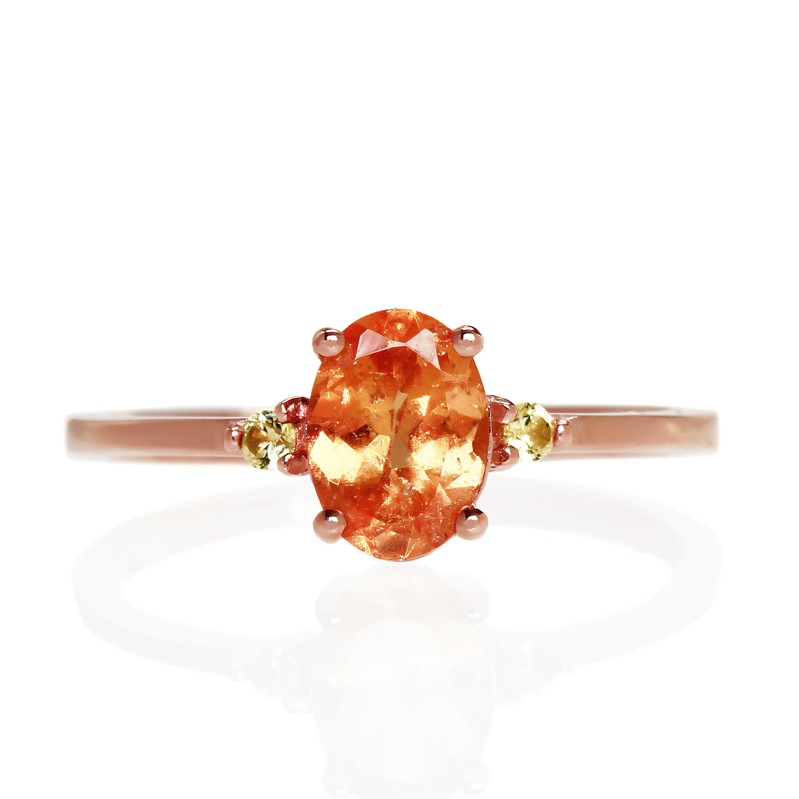 A product photo of a rose gold mandarin garnet ring sitting against a white background. The rose gold band is plain and smooth, and the centre oval-cut light bright orange centre stone is framed by a single yellow sapphire jewel on either side.
