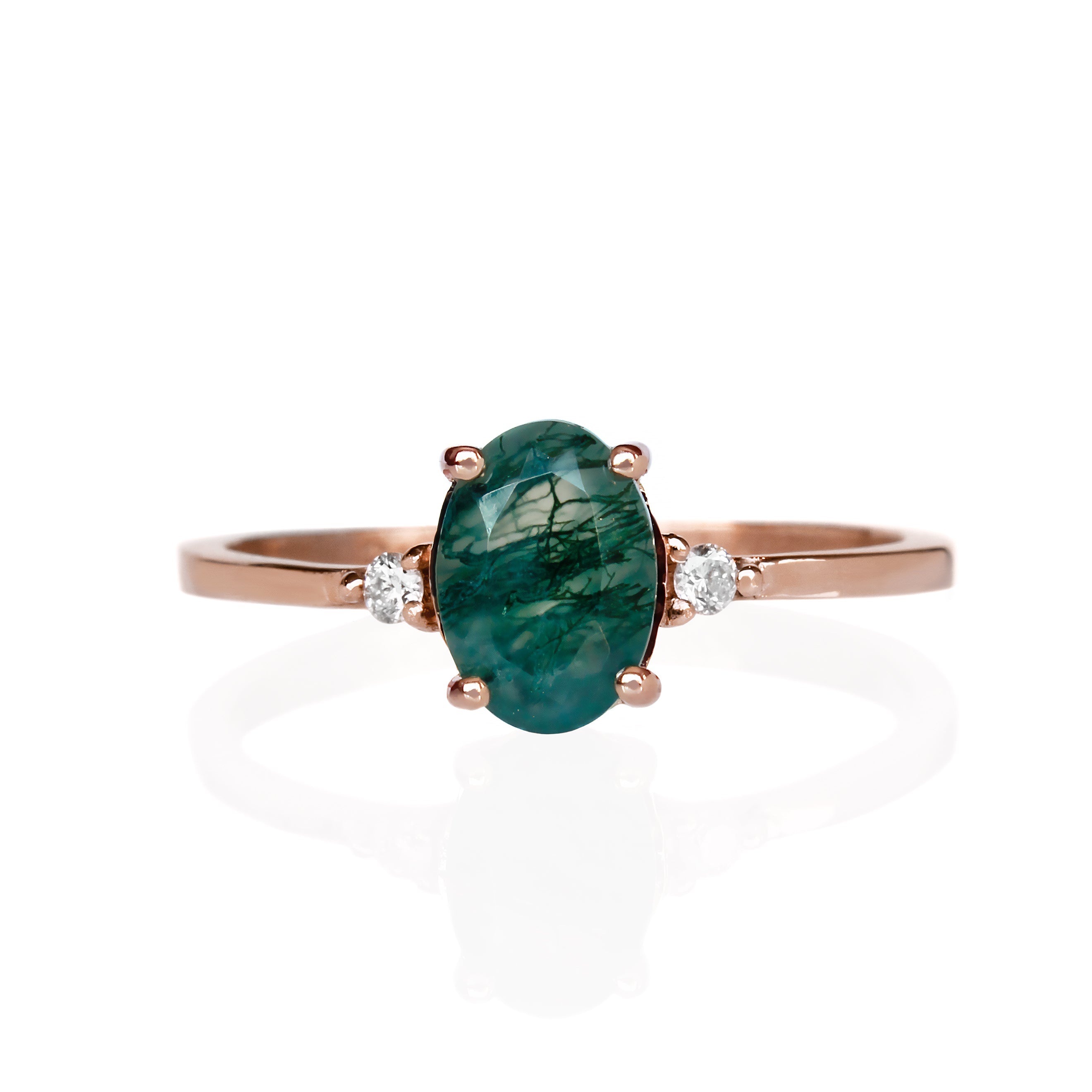 A product photo of a rose gold moss agate ring sitting against a white background. The rose gold band is plain and smooth, and the centre oval-cut deep green moss agate stone is framed by a single white diamond on either side. The moss agate stone is semi-opaque with stunning deep green tendrils of swirling dendritic inclusions.