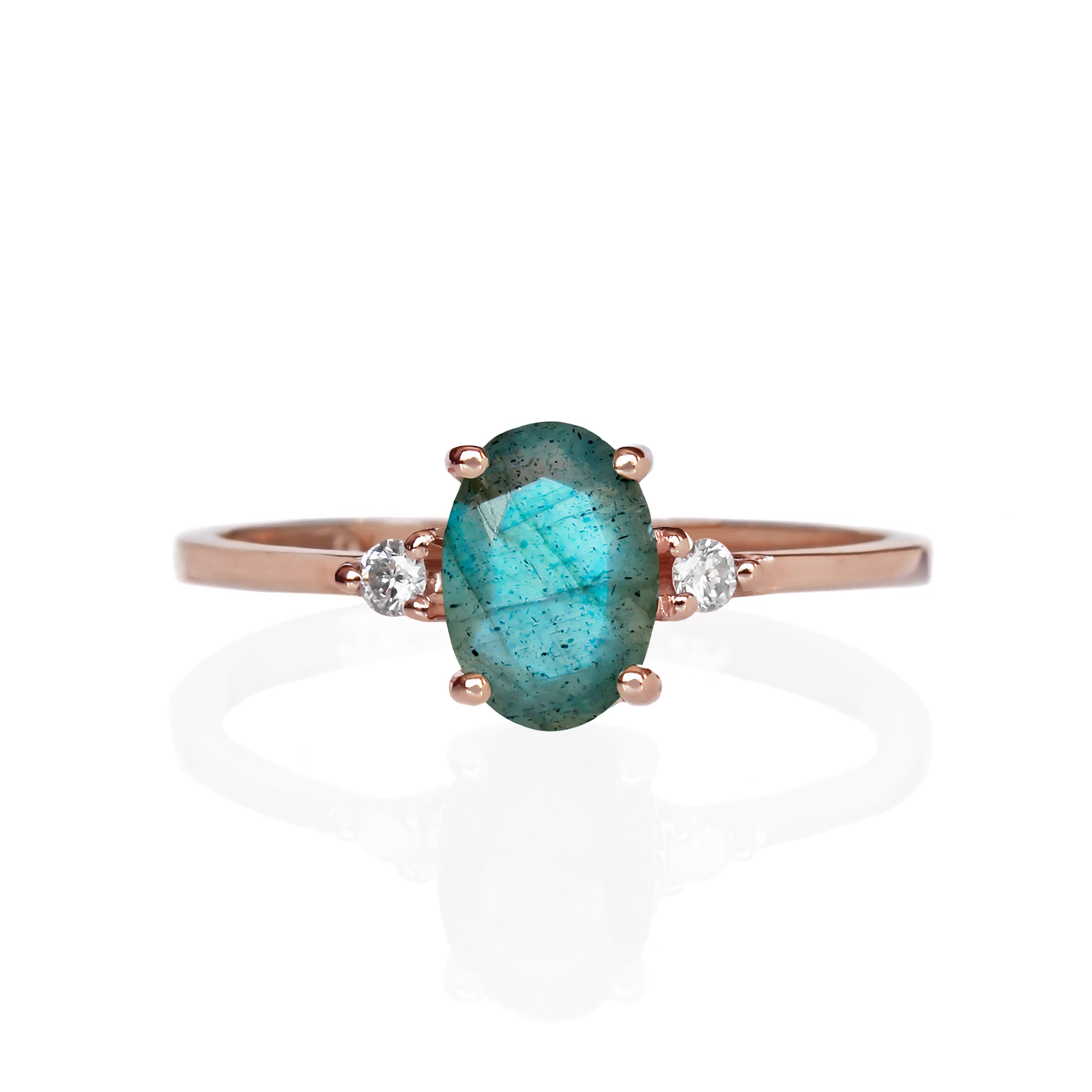 A product photo of a rose gold labradorite ring sitting against a white background. The rose gold band is plain and smooth, and the centre oval-cut colour-changing stone is framed by a single white diamond on either side.