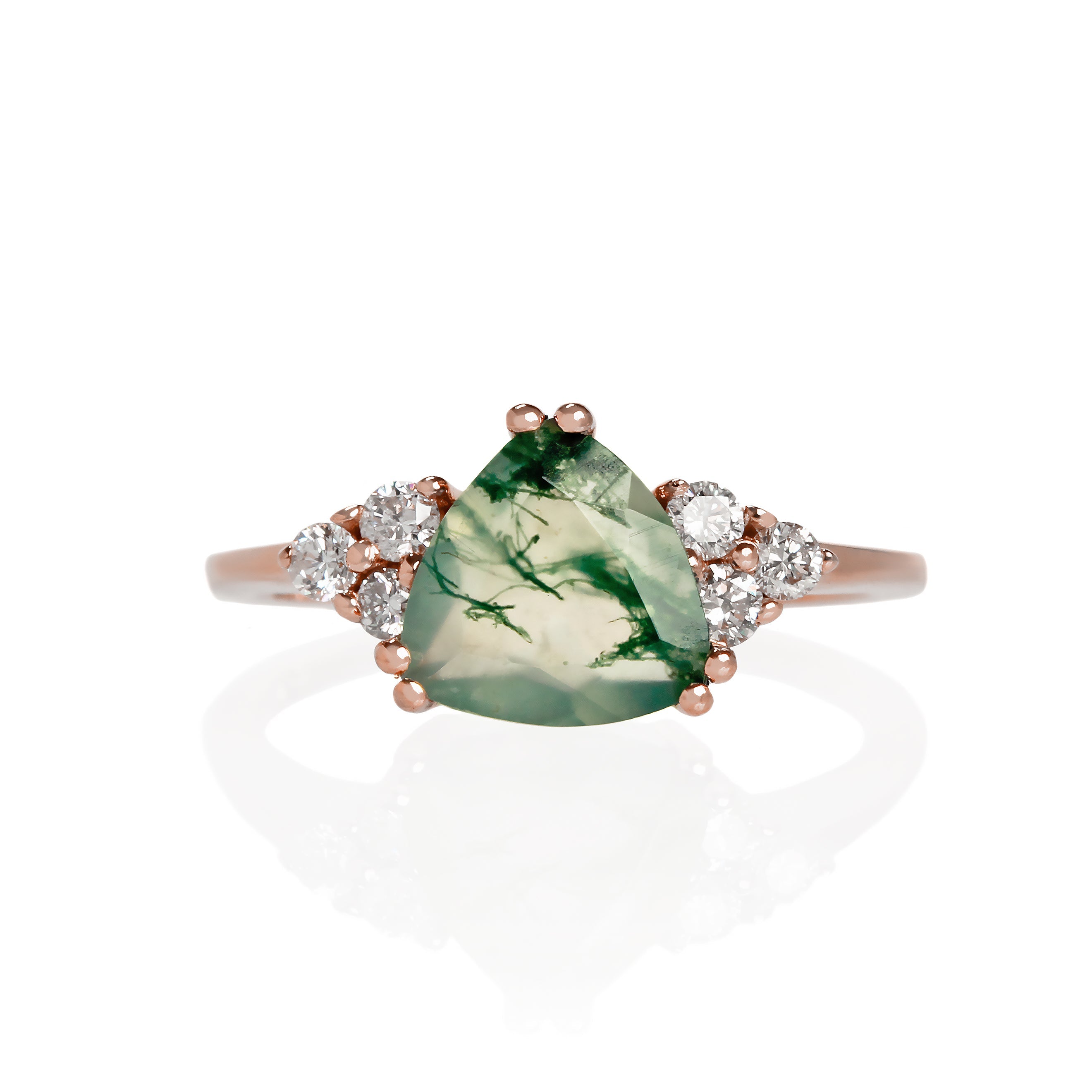 A product photo of a rose gold moss agate and diamond trio ring sitting on a white background. The trilliant-cut, naturally-included moss agate gemstone stands in stark contrast to the clusters of three classic white diamond stones on either side.